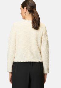 Cremfarbener Jacke mit texturierter Oberfläche, langen Ärmeln und Fransenverzierungen entlang des Saums und der Manschetten, kombiniert mit schwarzen Hosen. Rückansicht dargestellt.
