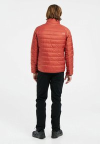 Giacca isolante rossa con colletto rialzato, design trapuntato e un piccolo logo sulla parte superiore della schiena, abbinata a pantaloni neri e calzature.