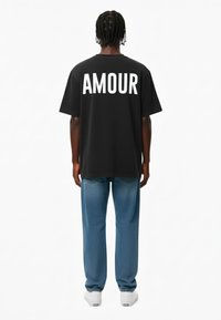Juoda per didelė t-shirt su baltais užrašu "AMOUR" ant nugaros, derinama su šviesiai mėlynomis džinsinėmis kelnėmis ir baltomis sportbačiais. Paprastas dizainas su kasdiene išvaizda.