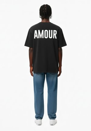 Zwarte oversized t-shirt met witte "AMOUR" tekst op de achterkant, gecombineerd met lichtblauwe denim jeans en witte sneakers. Eenvoudig ontwerp met een casual pasvorm.