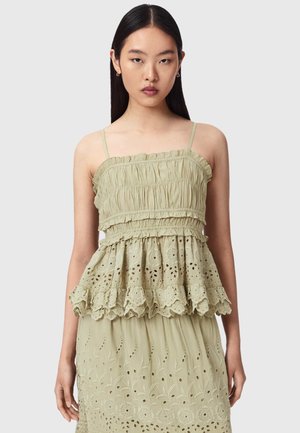 Femme aux cheveux noirs et raides portant un haut sans manches vert clair avec des volants et des détails en broderie anglaise, se tenant devant un fond uni.