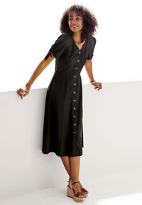 Schwarzes Hemdkleid mit kurzen Puffärmeln, V-Ausschnitt und tailliertem Schnitt. Trägt dazu braune Keilsandalen auf einem neutralen Untergrund.