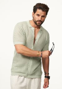 Strick-Shortsleeve-Cardigan in hellgrün mit einem Wabenmuster, ausgestattet mit einer durchgehenden Knopfleiste und gerippten Bündchen, getragen über hellen Hosen.