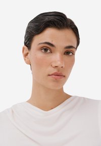 Detailní záběr na osobu s krátkými, slicked-back vlasy, oblečenou v bílém drapovaném topu. Má jemný make-up a hnědé oči, se světlou pletí.