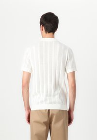 Suéter branco de malha de mangas curtas com um padrão texturizado em riscas e punhos canelados, combinado com calças bege.