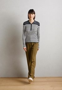 Chemise à rayures à manches longues avec un col foncé et une patte de boutonnage, associée à un pantalon vert olive et des baskets blanches sur un sol en bois.
