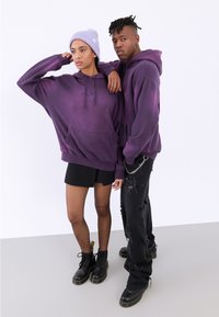 To modellene har på seg store, lilla hettegensere. Den kvinnelige modellen har på seg et svart miniskjørt og fiske-nett strømpebukser; den mannlige modellen har på seg slitte svarte jeans og støvler.