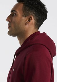 Bordeaux hoodie met ribbadige manchetten en zoom, voorzien van een voorste kangoeroe-pocket en een logo op de borst. Zacht fabric en een relaxte pasvorm.