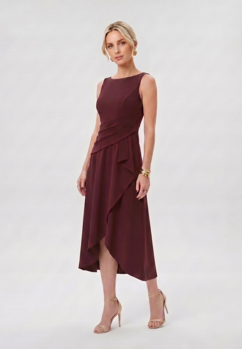 Femme aux cheveux blonds portant une robe sans manches bordeaux foncé avec un ourlet asymétrique et des sandales beiges à talons hauts, debout devant un fond uni.