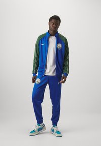 Blue tracksuit with green accents, featuring a basketball logo. White shirt underneath, paired with blue and white sneakers.