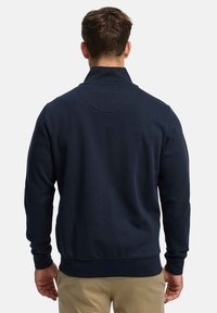 Sweat-shirt bleu marine avec col montant, poignets et ourlet côtelés. Présente une texture lisse et un empiècement arrondi à l'arrière.