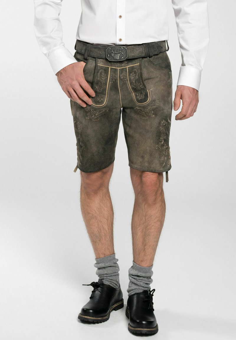 Braune Lederhosen mit bestickten Details und gelben Nähten, kombiniert mit grauen Socken und schwarzen Schuhen. Eine geknöpfte weiße Bluse ist sichtbar.