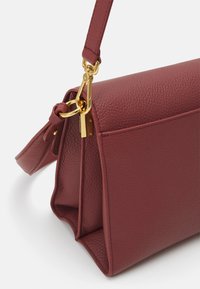 Μπορντό δερμάτινη τσάντα χειρός με υφή, χρυσές λεπτομέρειες και αφαιρούμενο λουρί. Διαθέτει δομημένο σχήμα και λεπτομέρειες με πλευρική τσέπη.