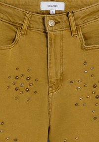 Pantalones de mezclilla color mostaza con un diseño ajustado, cierre de botón de latón y tachuelas decorativas dispuestas en varios tamaños en la parte frontal.