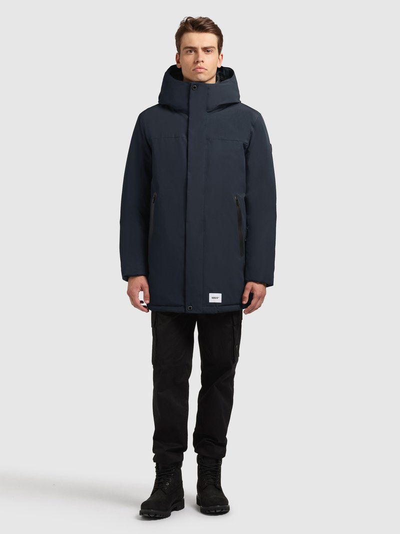 Marineblå vandtæt parka med høj krave, justerbar hætte, frontlynlås og sidelommer med lynlås. Kombineret med sorte cargobukser og støvler.