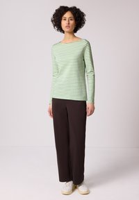 Mujer con cabello rizado que lleva una camisa de manga larga a rayas verdes claras y blancas, pantalones anchos oscuros y zapatillas blancas de pie.