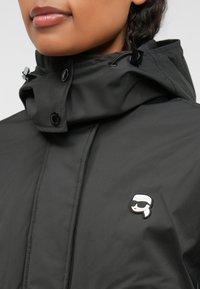 Juodas vandeniui atsparus švarkas su aukštu apykakle, turintis mygtukų užsegimus ir reguliuojamą gobtuvą. Išskirtinis siuvinėjimo logotipas ant krūtinės.