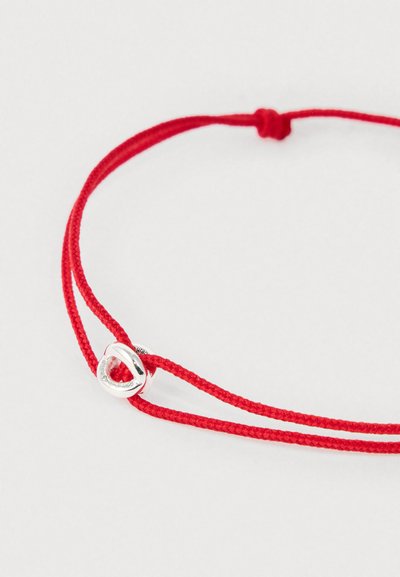 Braccialetto in corda intrecciata rossa con nodo regolabile e piccolo charm a forma di cuore in argento su sfondo bianco.