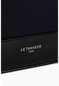 Schwarzes Leder-Akzent, geripptes navy-blaues Gewebe und geprägte Markenbezeichnung "LE TANNEUR 1898" unten rechts. Glatte Textur und klare Linien.