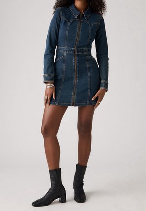 Femme portant une robe en jean bleu foncé ajustée avec fermeture éclair, manches longues et bottines noires, debout devant un fond clair uni.