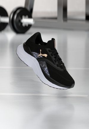 Czarny sportowy sneaker z białą podeszwą i złotym logo Puma, pokazany w powietrzu na podłodze siłowni z rozmytymi ciężarami w tle.