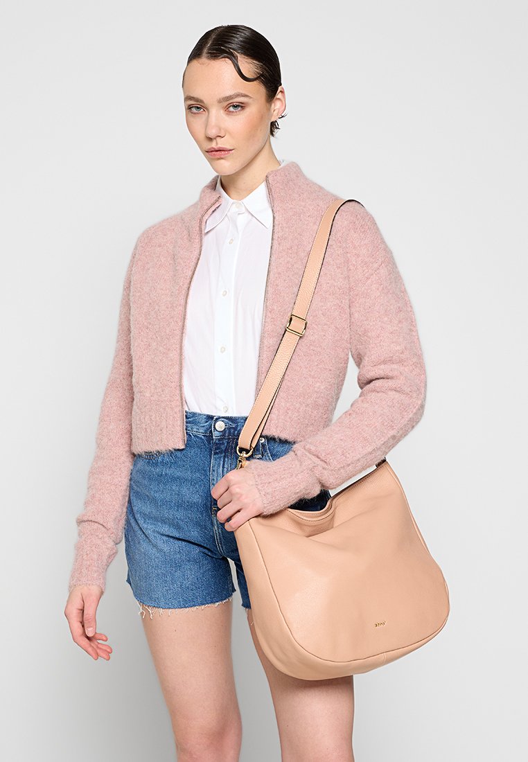 Rosa gestrickter, kurzer Pullover, weißes Shirt, Jeanshorts, hält eine große beigefarbene Ledertasche mit glatter Textur und goldenen Hardware-Details.