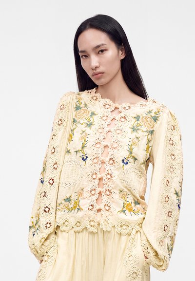 Woman with long dark hair wears cream lace blouse with floral embroidery and puff sleeves against plain light background.