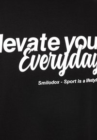 Sort bomulds T-shirt med fed hvid tekst, der står "Elevér din hverdag" og mindre tekst "Smilodox - Sport er en livsstil."