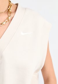 Beige Nike ärmlös topp med djup V-ringning, ribbad kant och en liten vit logotyp på bröstet; mjukt bomullsblandat tyg.
