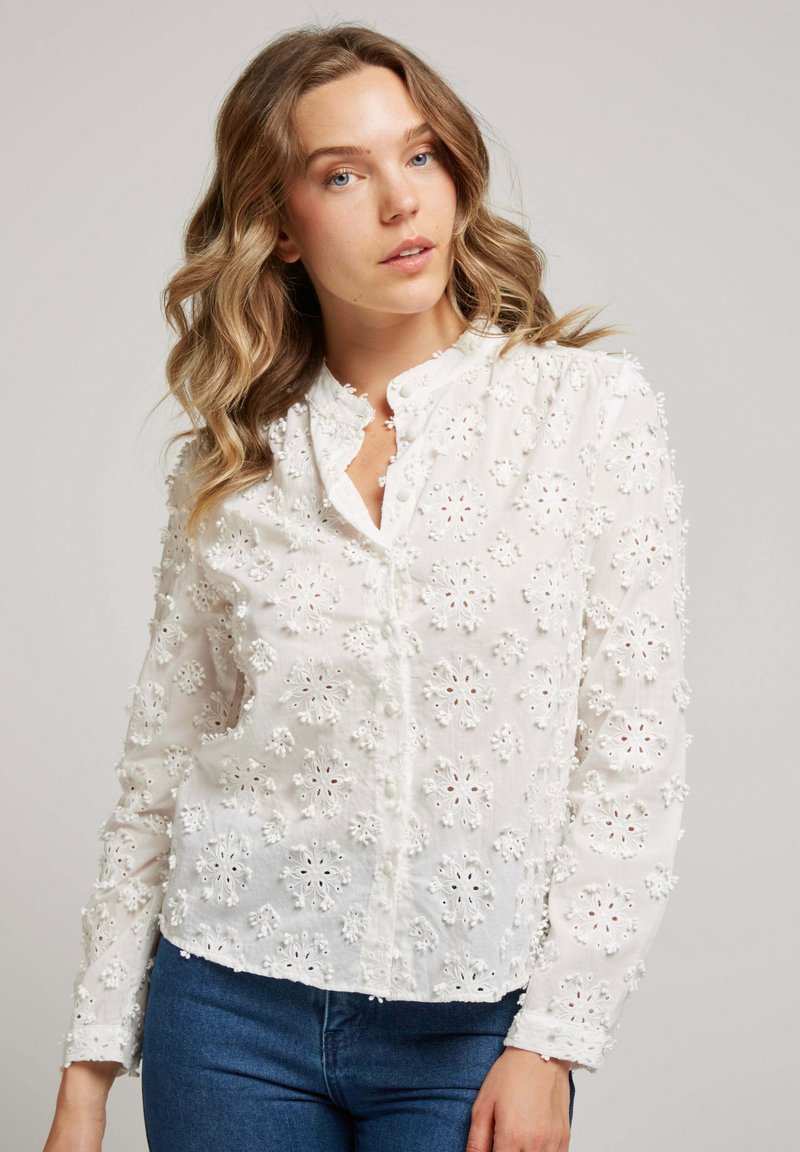 Blouse blanche avec broderie florale, dotée d'un col rond, d'une fermeture à boutons et d'un design court. Tissu doux et texturé avec des broderies complexes.