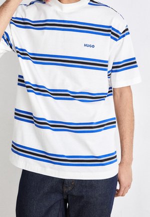 Hvid T-shirt med vandrette blå og sorte striber, "HUGO"-logo på brystet, båret med mørkeblå jeans, persons hånd nær ansigtet.