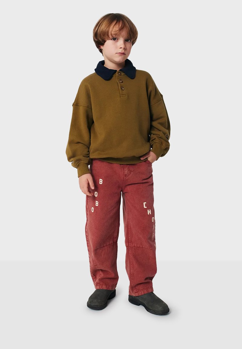 Sweat-shirt vert olive avec col marine, trois boutons, coupe ample. Pantalon rouge avec texte blanc « BOBO CHO », design à jambe droite, porté avec des chaussures noires.