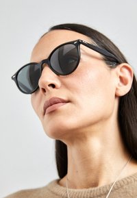 Ochelari de soare negri cu formă de pisică, cu lentile întunecate, accente din metal și o finisare mată. Rama este subțire și unghiulară, ceea ce subliniază designul modern.