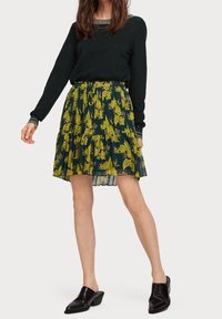 Jupe plissée florale en vert foncé avec des motifs de fleurs jaunes, en matériau léger, design superposé, associée à un haut et des chaussures noirs.