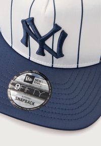 Námornícky modrá šiltovka snapback s bielymi prúžkami. Obsahuje vyšívané logo NY a striebornú značkovú nálepku na šilt. A-rámový dizajn.