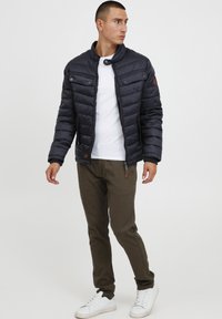 Marineblå pufferjakke med høy krage, glidelås, frontlommer og brune skinnakenter. Kombinert med olivenfargede bukser og hvite joggesko.