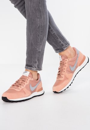 Henkilö, jolla on harmaat farkut ja vaaleanpunaiset Nike-tossut harmaalla swoosh-logolla, seisoo valkoista taustaa vasten.