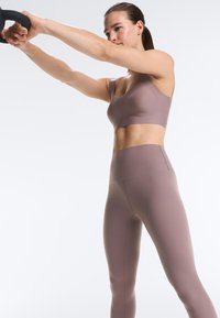 Kobieta w mauve sportowym biustonoszu i legginsach ćwiczy z czarnym pierścieniem fitness, wyciągając ramiona do przodu na białym tle.