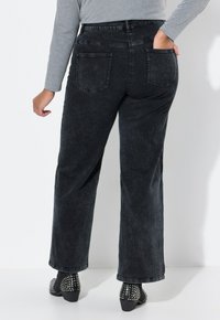 Jeans en denim noir à jambes larges avec poches arrière, présentant une texture délavée. Associés à des bottes à talons cloutées, mettant en valeur un look soigné.