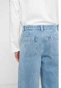 Πίσω όψη ατόμου που φοράει ανοιχτό μπλε τζιν με μπαλώματα στις τσέπες και λευκό πουκάμισο με μακριά μανίκια κεντημένο μέσα.