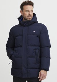 Marineblå pufferjakke med hette, med kviltet design, frontglidelås, sidelommer og logodetaljer. Glatt tekstur, isolert materiale.