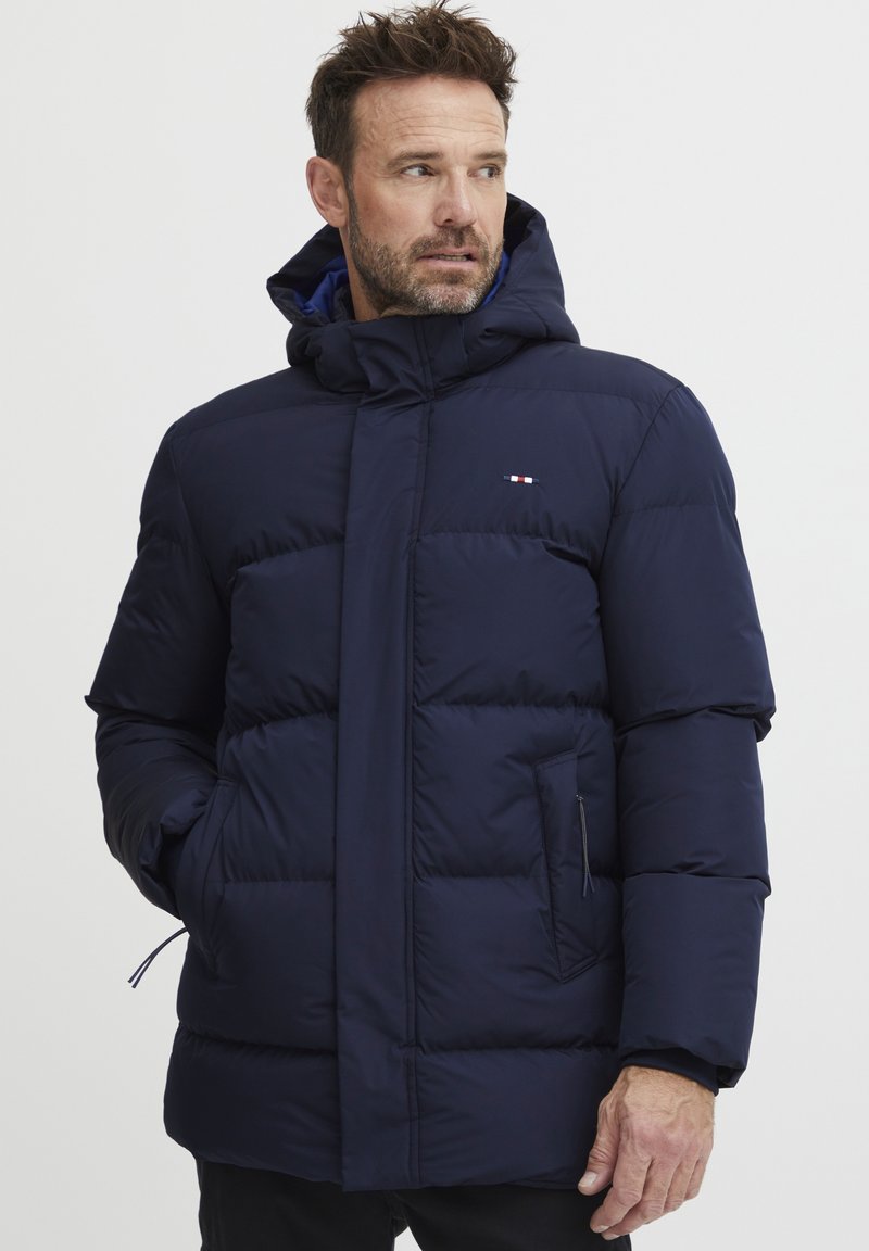 Marineblå pufferjakke med hette, med kviltet design, frontglidelås, sidelommer og logodetaljer. Glatt tekstur, isolert materiale.