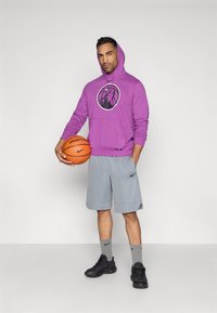 Sweatshirt roxa com capuz e logotipo preto, calções de desporto cinzentos, sapatilhas pretas e meias cinzentas. Segurando uma bola de basquete laranja.