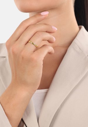 Mano con unghie rosa chiaro che indossa un semplice anello d'oro, posizionata vicino al collo e indossa un blazer beige su una maglietta bianca.