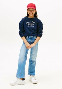 Sudadera navy con logo blanco, de manga larga, combinada con jeans rectos de un azul claro y zapatillas blancas. El modelo lleva una gorra roja.