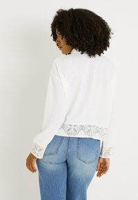 Suéter blanco de mangas largas con adornos de encaje en el dobladillo y los puños, combinado con jeans azules. La tela parece suave y tiene un diseño casual.