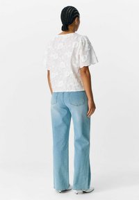 Hvid broderet bluse med korte ærmer, parret med lyseblå lige ben jeans. Blusen har blomster mønstre og en bølget kant.