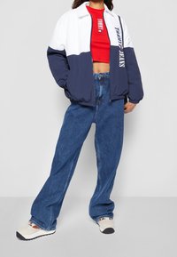 Navy and white jacket with a zip closure, paired with a red cropped shirt and loose blue jeans. Shoes feature a cream base with black accents.