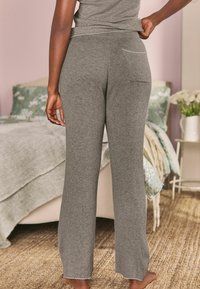 Šedé žebrované pletené kalhoty s volným střihem, s kapsou vzadu a vroubkovanými lemy, zobrazené na pozadí jemně zařízené ložnice.