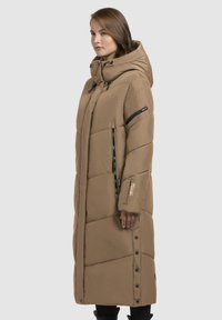 Garš brūns pūkains mētelis ar kapuci, kuram ir izšūts raksts, melna rāvējslēdzēju apdare un spiedpogu aizdare sānos.